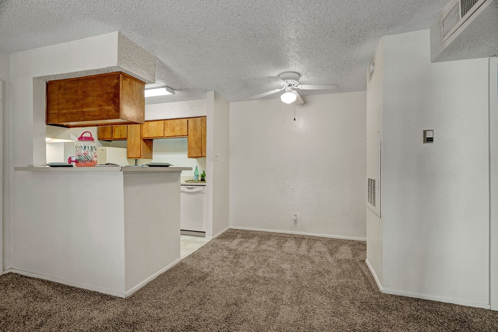 Cozy open-plan apartment here at The Bradford Apartment Homes with white walls, brown carpet, and a small kitchen with wooden cabinets. A ceiling fan adds a calm atmosphere.