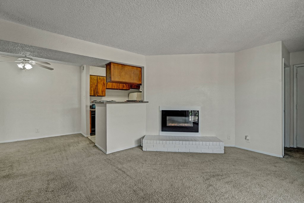 Living room here at The Bradford Apartment Homes with beige carpet, white walls, and a white brick fireplace. Open kitchen with wooden cabinets and a ceiling fan in the corner. Cozy ambiance.