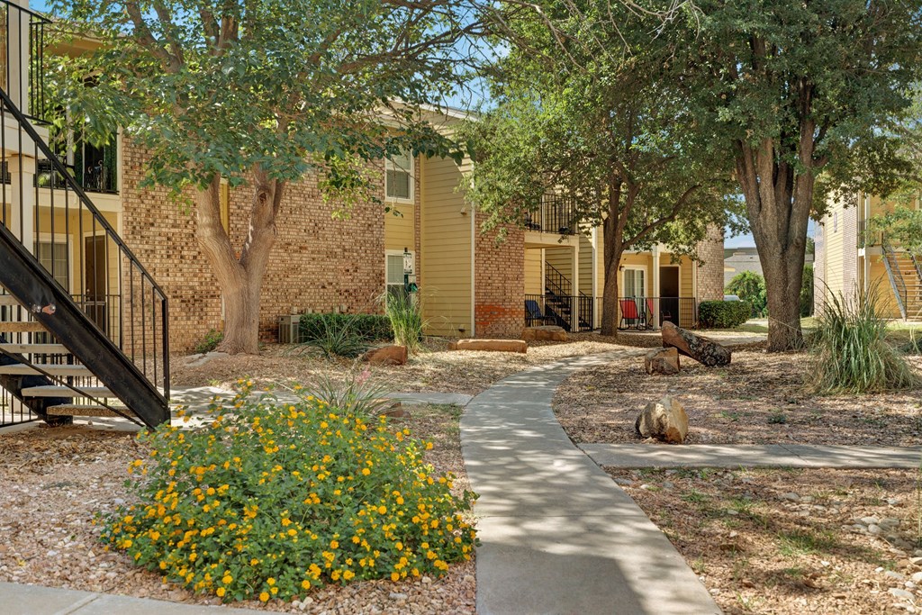 Curved path here at The Bradford Apartment Homes leads to brick apartments, shaded by large trees. Yellow flowers and shrubs line the walkway, creating a peaceful, welcoming atmosphere.