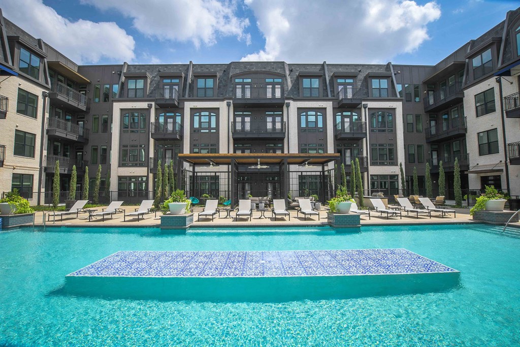 Luxurious The Crosby at The Brickyard Apartment Homes complex with a large, inviting swimming pool in the foreground. Lounge chairs and greenery surround the pool. Clear, sunny day.