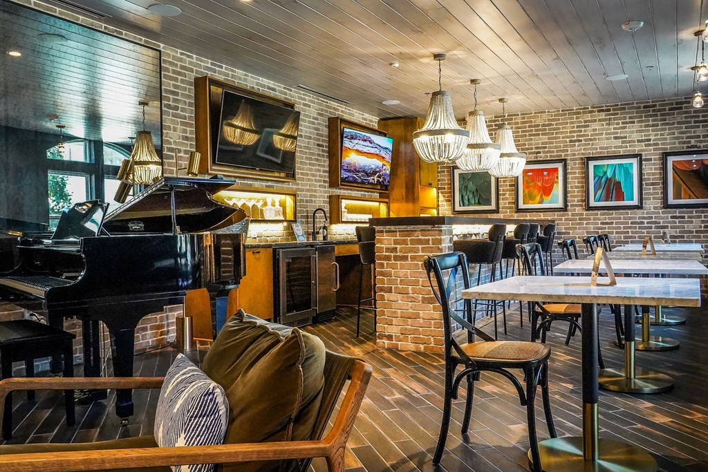 Chic lounge here at The Crosby at the Brickyard Apartment Homes with a grand piano, brick walls, and chandeliers. Marble tables and bar stools create an elegant, relaxed atmosphere. Vibrant paintings add warmth.