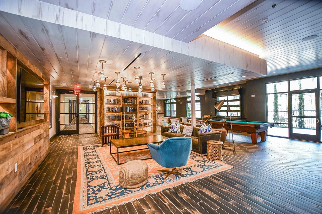 Stylish lounge here at The Crosby at the Brickyard Apartment Homes with modern decor, featuring wooden floor, blue and orange rug, blue chair, brown sofas, pool table, and large windows. Cozy ambiance.