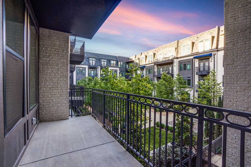View from a modern balcony here at The Crosby at the Brickyard Apartment Homes overlooking a courtyard with lush green trees and stylish apartment buildings. The sky is vivid with hues of pink and blue.