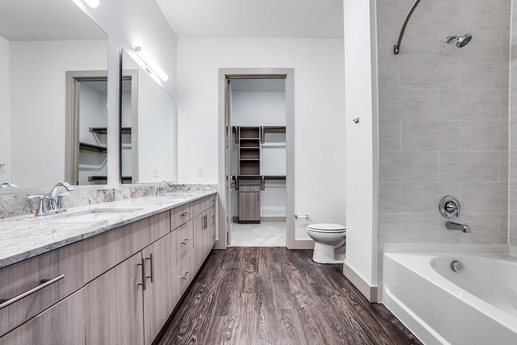 Modern bathroom here at The Crosby at the Brickyard Apartment Homes with wood floors, dual marble sinks, large mirror, and a tiled bathtub. Calm, neutral tones create a sleek, relaxing space.