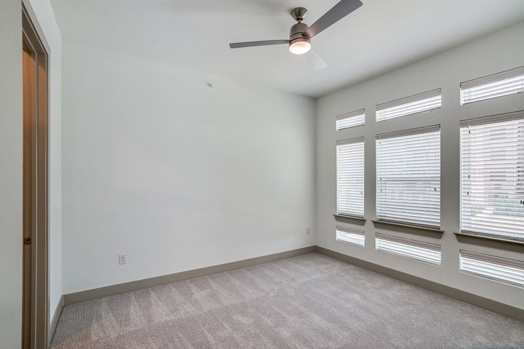 Empty bedroom here at The Crosby at the Brickyard Apartment Homes with gray carpet, white walls, and a ceiling fan. Bright light streams through multiple narrow windows with closed blinds, creating a calm ambiance.