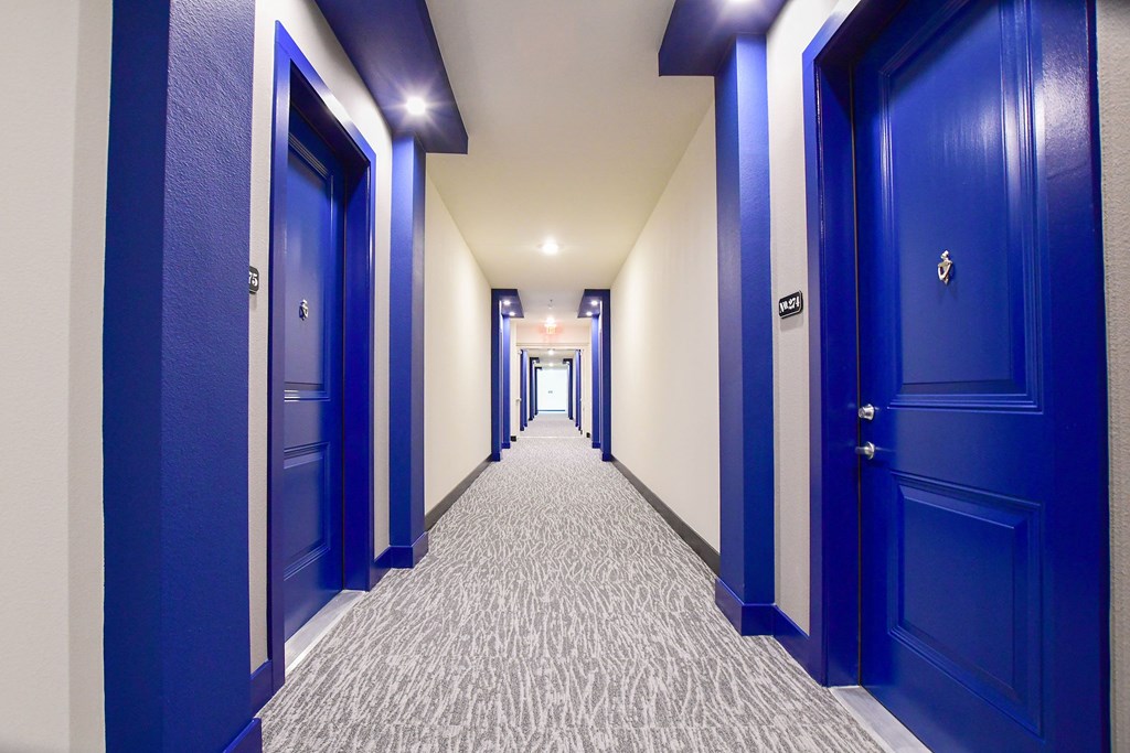 A long hallway here at The Crosby at the Brickyard Apartment Homes with textured carpet leads to a bright exit. Blue doors and frames line the walls, creating a symmetrical and modern feel.