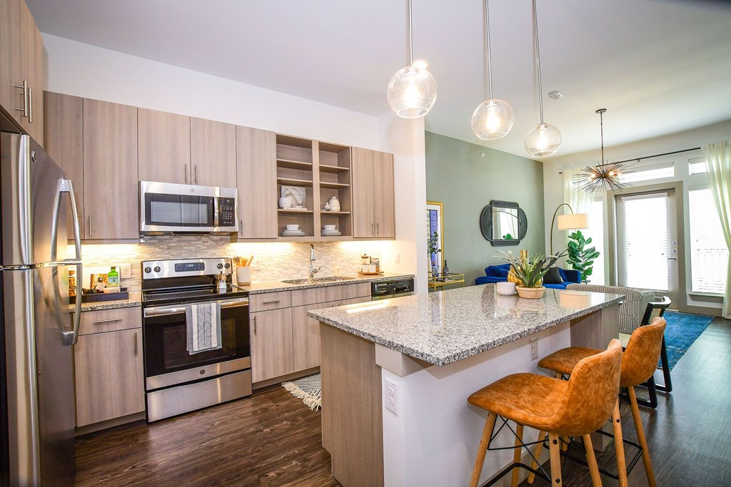 Modern kitchen here at The Crosby at the Brickyard Apartment Homes with light wood cabinets, stainless steel appliances, and granite island. Two leather stools, hanging lights, and a cozy living area beyond.