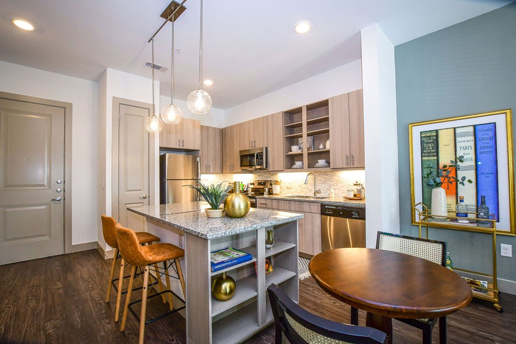 Modern kitchen here at The Crosby at the Brickyard Apartment Homes with light wood cabinets, granite island, and three pendant lights. Orange barstools, framed art, and plants add warmth and style.
