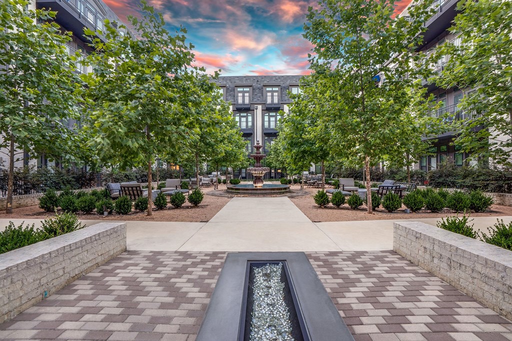 Peaceful courtyard here at The Crosby at the Brickyard Apartment Homes with trees, a fountain centerpiece, and benches. Paved path leads to a modern building under a vibrant, colorful sunset sky.