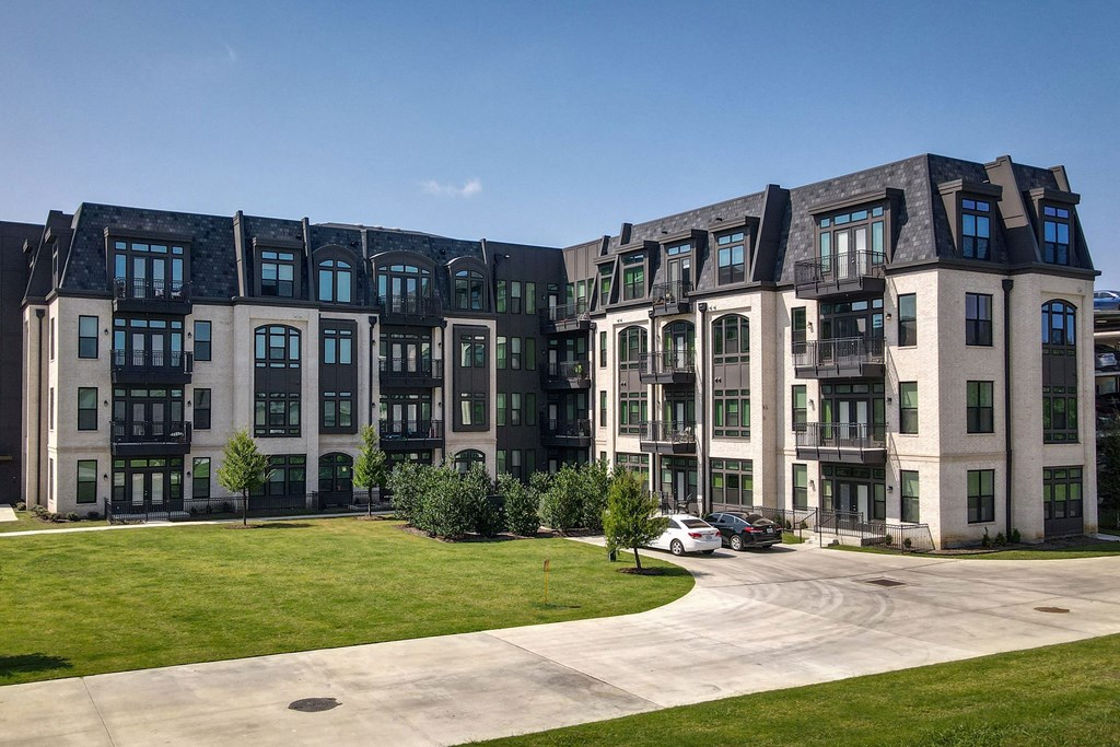The Crosby at The Brickyard Apartment Homes building with multiple floors and arched windows under a clear blue sky. There's a curved driveway, lush green lawn, and parked cars.