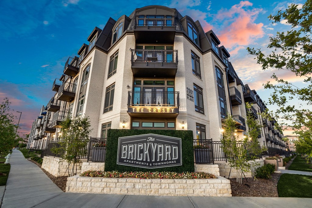 Four-story brick apartment building with balconies, labeled "The Crosby" and "The Brickyard Apartments & Townhomes." Evening sky glows with pink and blue hues.