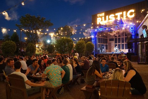 A lively outdoor restaurant at night with people dining under string lights, a warm, festive atmosphere, and the sign "RUST" glowing in the background.