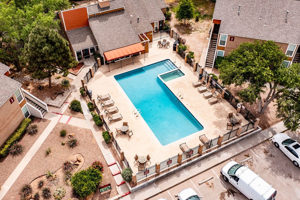 Aerial view of a rectangular outdoor swimming pool here at Trinity Place Apartment Homes surrounded by lounge chairs and a fence, nestled between modern apartment buildings with trees nearby.