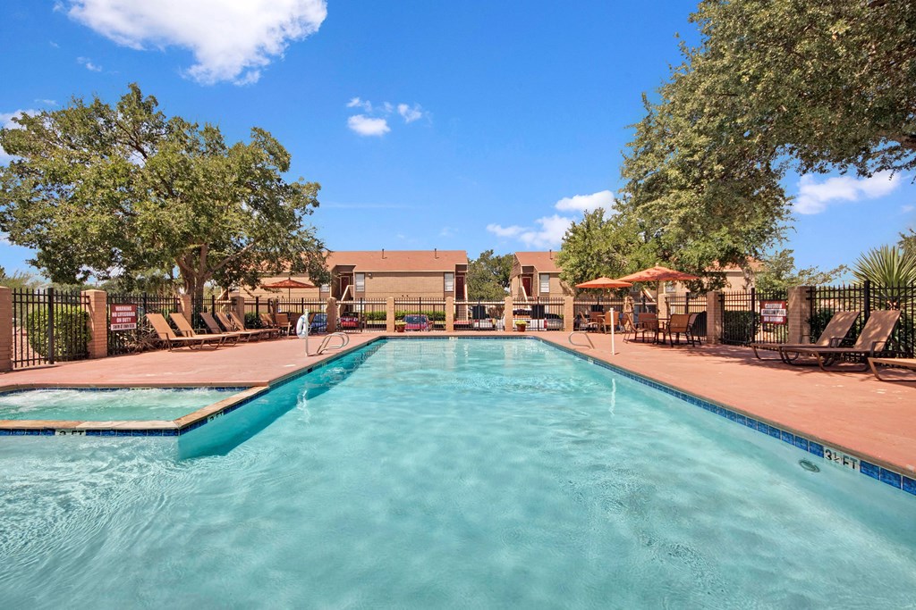 A tranquil outdoor pool here at Trinity Place Apartment Homes under a bright blue sky, surrounded by trees, lounge chairs, and orange parasols, creating a relaxing, sunny atmosphere.
