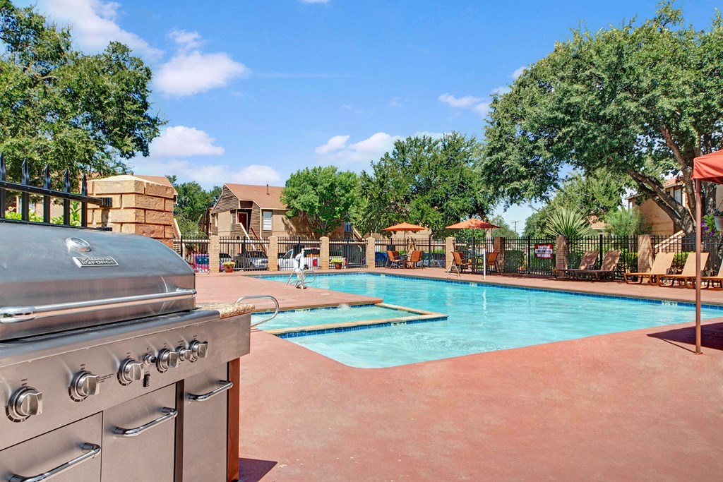 Outdoor pool area here at Trinity Place Apartment Homes with a stainless steel grill in the foreground, surrounded by red brick patio, lounge chairs, and umbrellas under a blue sky.