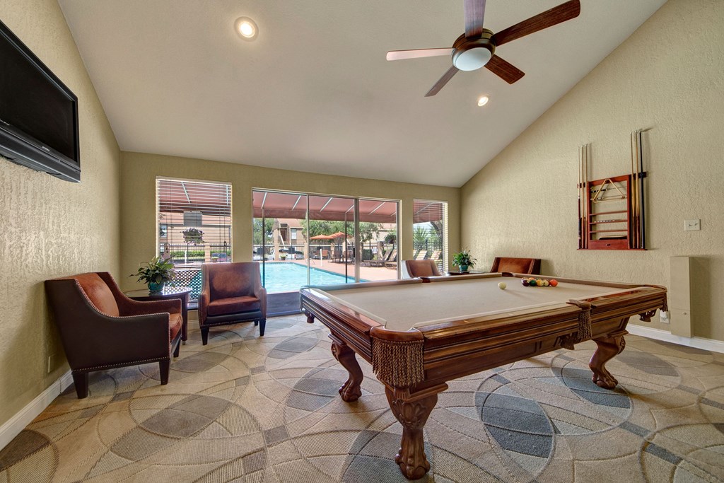 A cozy room here at Trinity Place Apartment Homes with a pool table, elegant chairs, and large windows overlooking a sunny outdoor pool area. Walls are beige, and a ceiling fan hangs above.