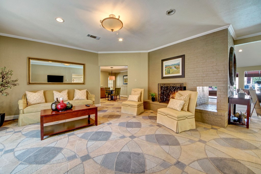 Spacious living room here at Trinity Place Apartment Homes with beige walls, a patterned carpet, and modern lighting. Includes a cream sofa, two armchairs, red coffee table, and wall art, exuding comfort.