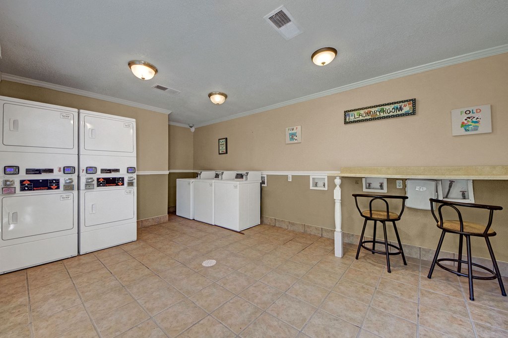 Laundry room here at Trinity Place Apartment Homes with beige walls and tiled floor. Features two stacked dryers, three washers, and a counter with two bar stools. Bright and clean atmosphere.