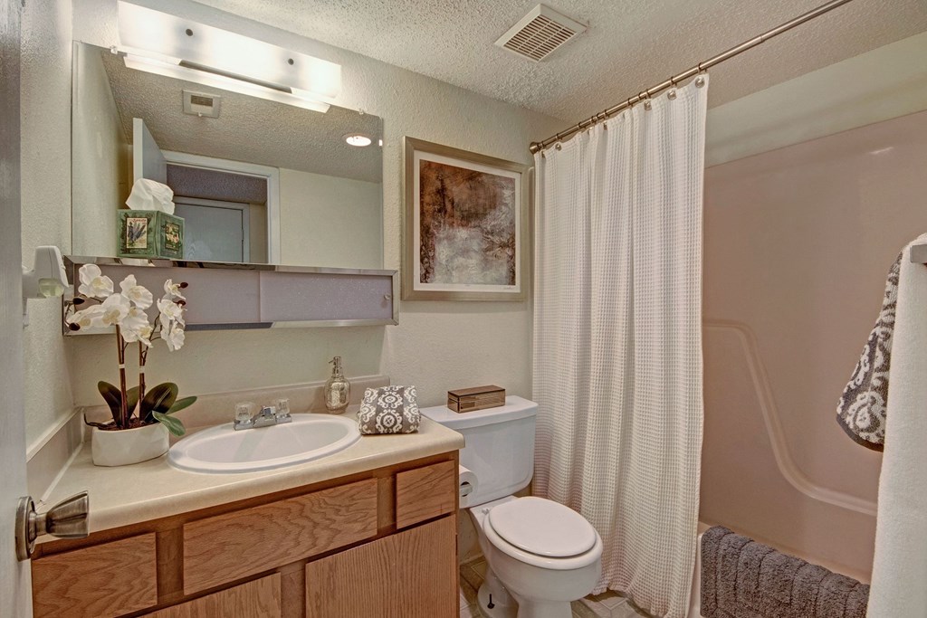 Small, warmly-lit bathroom here at Trinity Place Apartment Homes with a wooden vanity, decorated with orchids, soap dispenser, and a patterned tissue box. A framed abstract art piece hangs above a white toilet, next to a shower with a curtain.