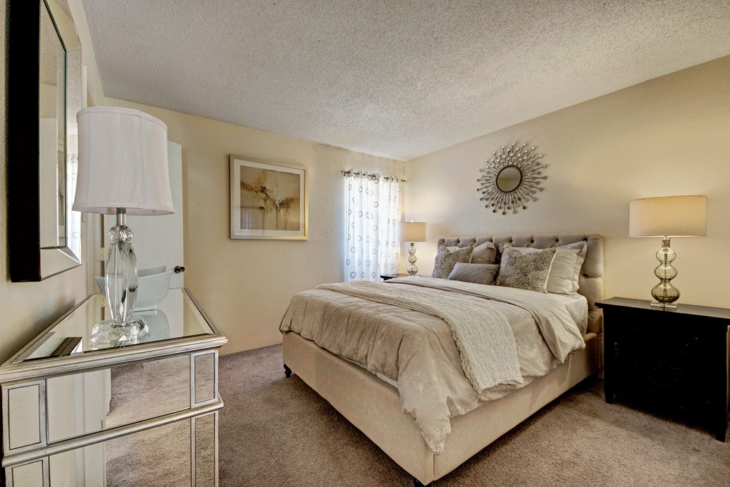 A cozy bedroom here at Trinity Place Apartment Homes with a beige color scheme, featuring a tufted bed, decorative pillows, mirrored dresser, and soft lighting from lamps and window.