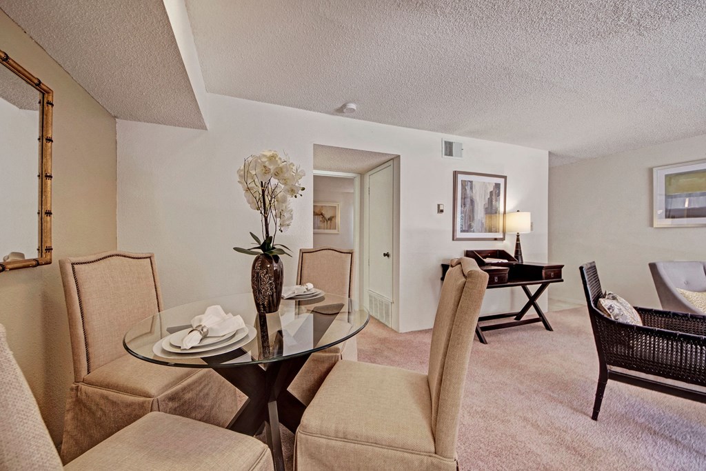A cozy living room here at Trinity Place Apartment Homes features a glass dining table with four padded chairs and an orchid centerpiece. A writing desk with a lamp is in the back, adding warmth.