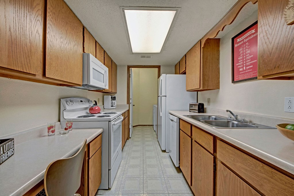Narrow kitchen here at Trinity Place Apartment Homes with wood cabinets and white appliances, including a stove, microwave, and fridge. Bright lighting and a cozy, clean ambiance.