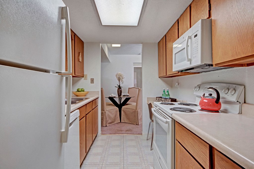 Narrow kitchen here at Trinity Place Apartment Homes with wooden cabinets, white appliances, and a red kettle on the stove. In the background, a dining table with flowers is visible.