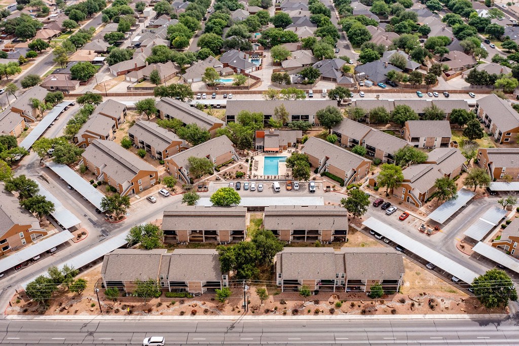 Aerial view of Trinity Place Apartment Homes with several brown buildings arranged around a central courtyard featuring a swimming pool. Surrounding green trees and nearby homes create a suburban atmosphere.