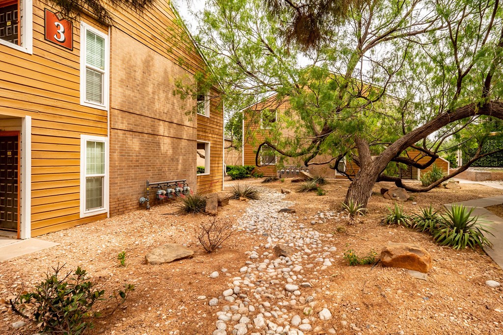 Apartment complex with tan brick buildings and a rocky, landscaped courtyard featuring a large, bent tree here at Trinity Place Apartment Homes. The scene is calm and inviting.
