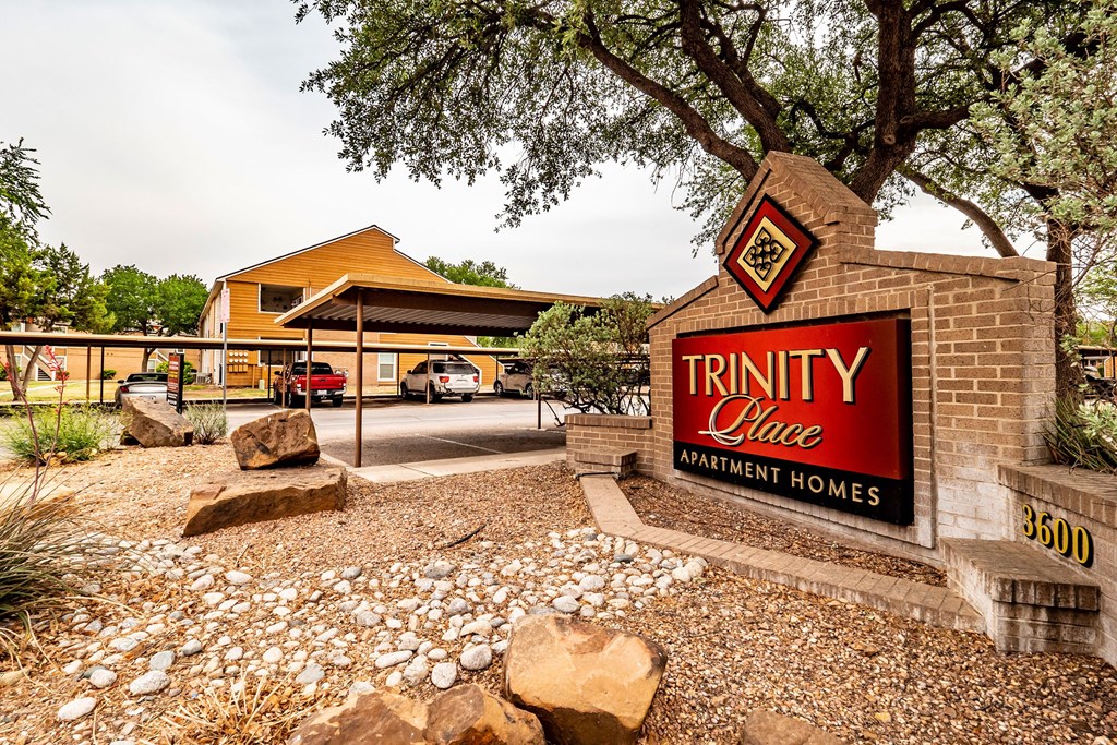 Trinity Place Apartment Homes entrance with a large, red sign. Surrounding landscape features rocks, desert plants, and a tree, creating a welcoming atmosphere.
