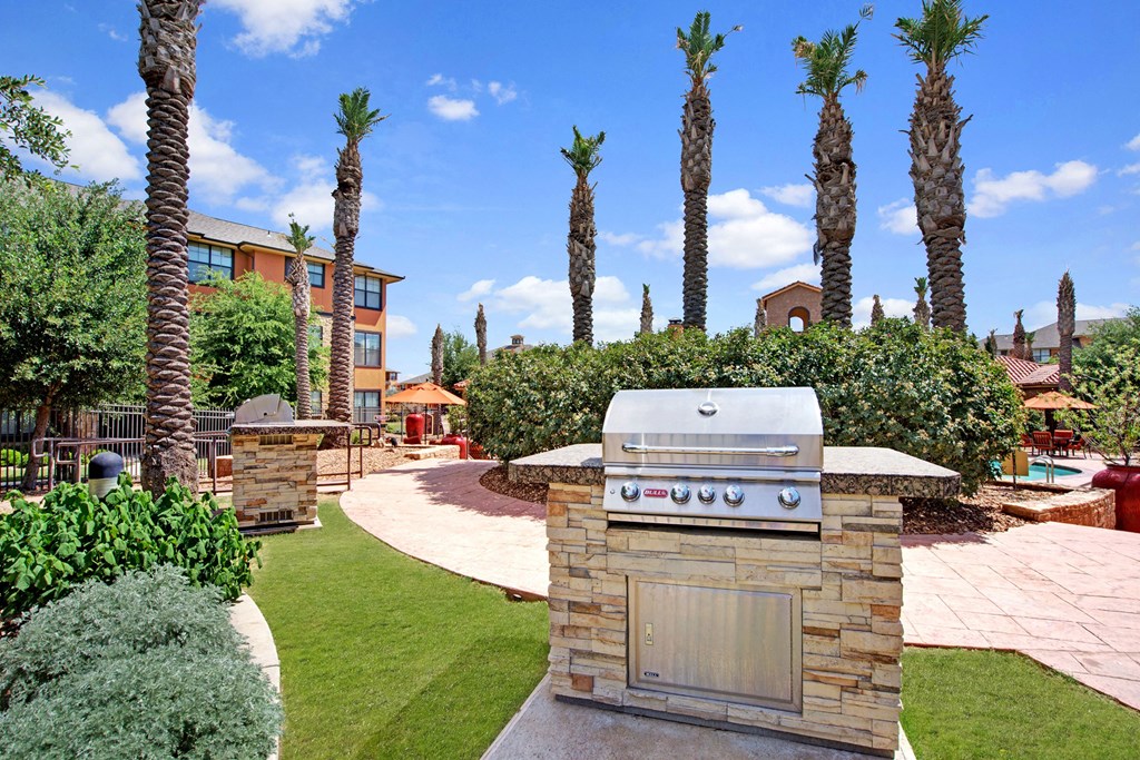 Outdoor scene here at Tuscany at Faudree Apartment Homes with a stainless steel barbecue grill on a stone island, surrounded by palm trees, green lawn, and a path. Apartment buildings and clear blue sky in the background convey a relaxed, sunny atmosphere.