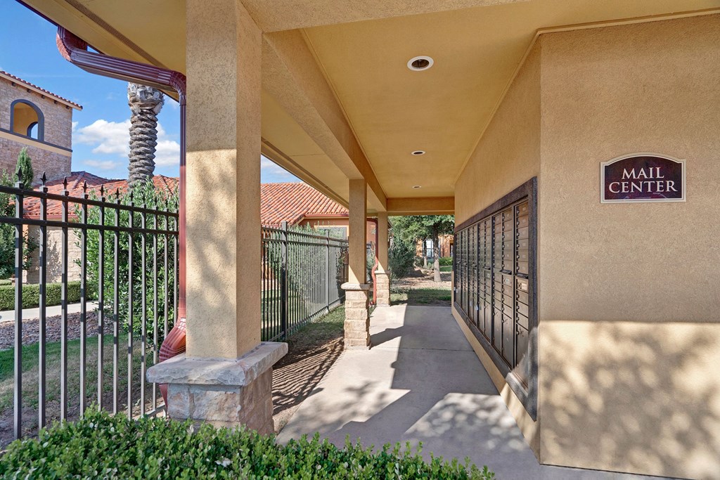 Covered walkway leading to a mail center here at Tuscany at Faudree Apartment Homes with beige stucco walls and a row of mailboxes. Sunlit shadows and greenery create a calm atmosphere.