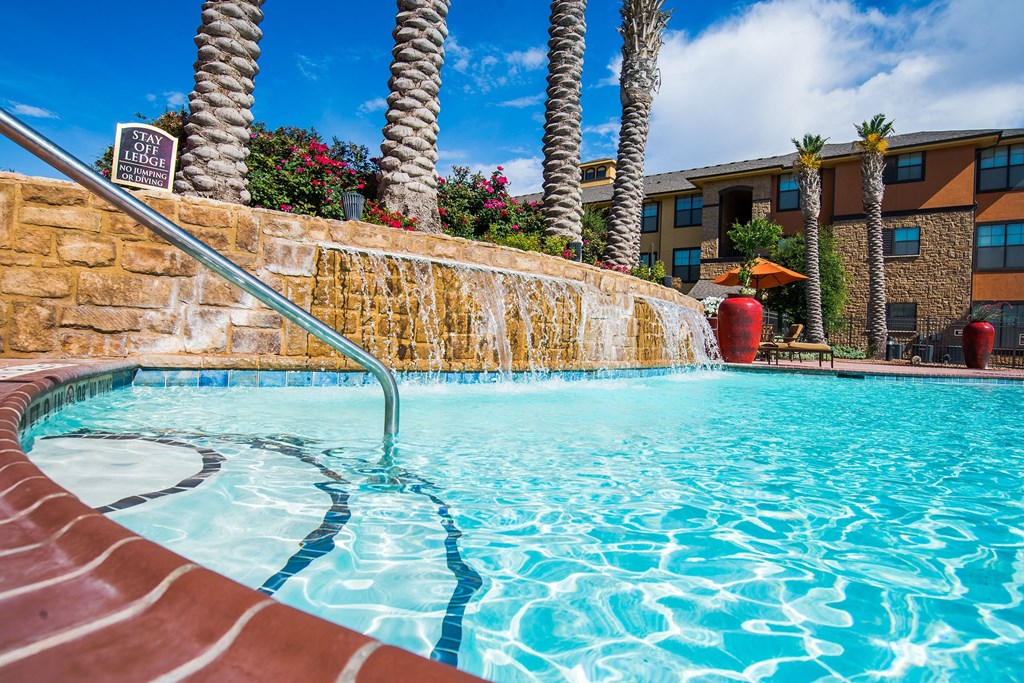 Sunny pool scene here at Tuscany at Faudree Apartment Homes with a rock waterfall, surrounded by palm trees and colorful flowers. Adjacent hotel buildings give a relaxing resort vibe.