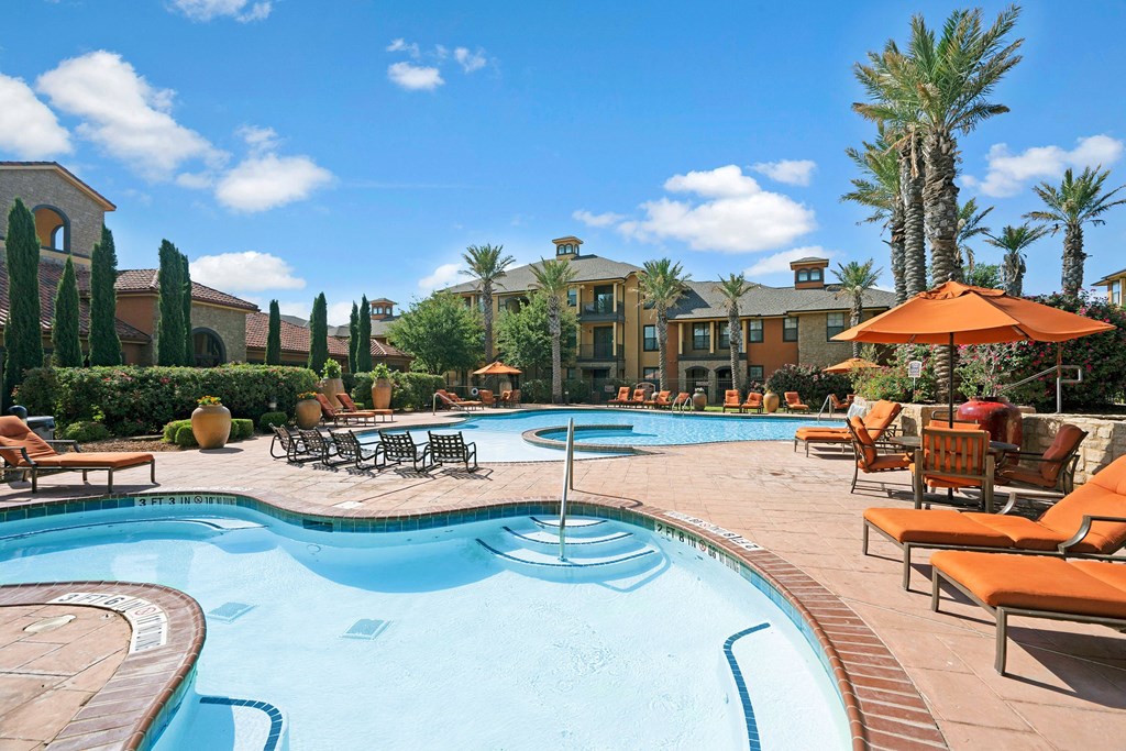 Luxurious outdoor pool area here at Tuscany at Faudree Apartment Homes with lounge chairs and umbrellas. Palm trees and vibrant greenery surround the space, under a clear, blue sky.