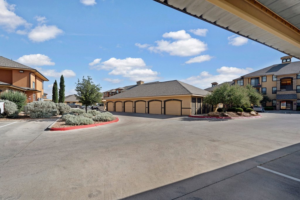 Sunny day here at Tuscany at Faudree Apartment Homes with beige buildings, garages, and neatly trimmed bushes. Blue sky with fluffy clouds adds a calm, inviting atmosphere.