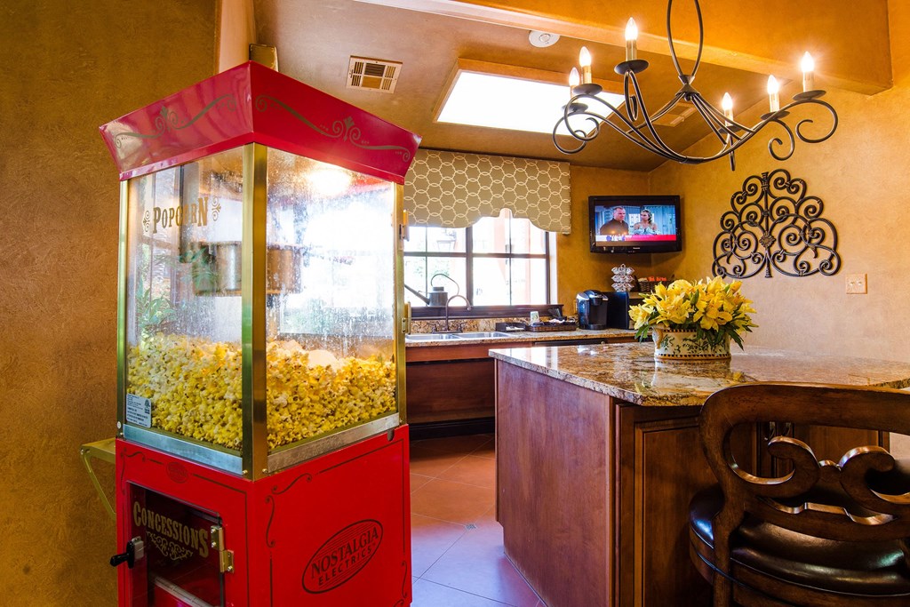 A cozy kitchen here at Tuscany at Faudree Apartment Homes features a red popcorn machine filled with popcorn beside a granite countertop with flowers. Warm lighting and a wall-mounted TV add comfort.