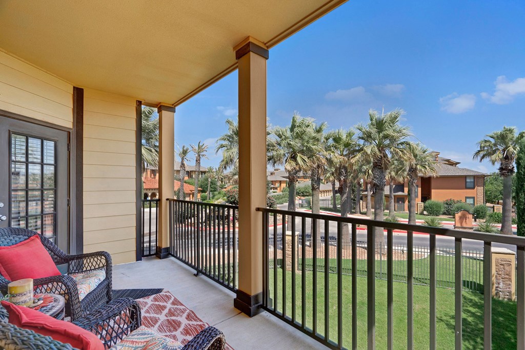 Covered balcony here at Tuscany at Faudree Apartment Homes with black railing, wicker chairs, and red cushions overlooks a sunny landscape of palm trees and buildings under a clear blue sky.
