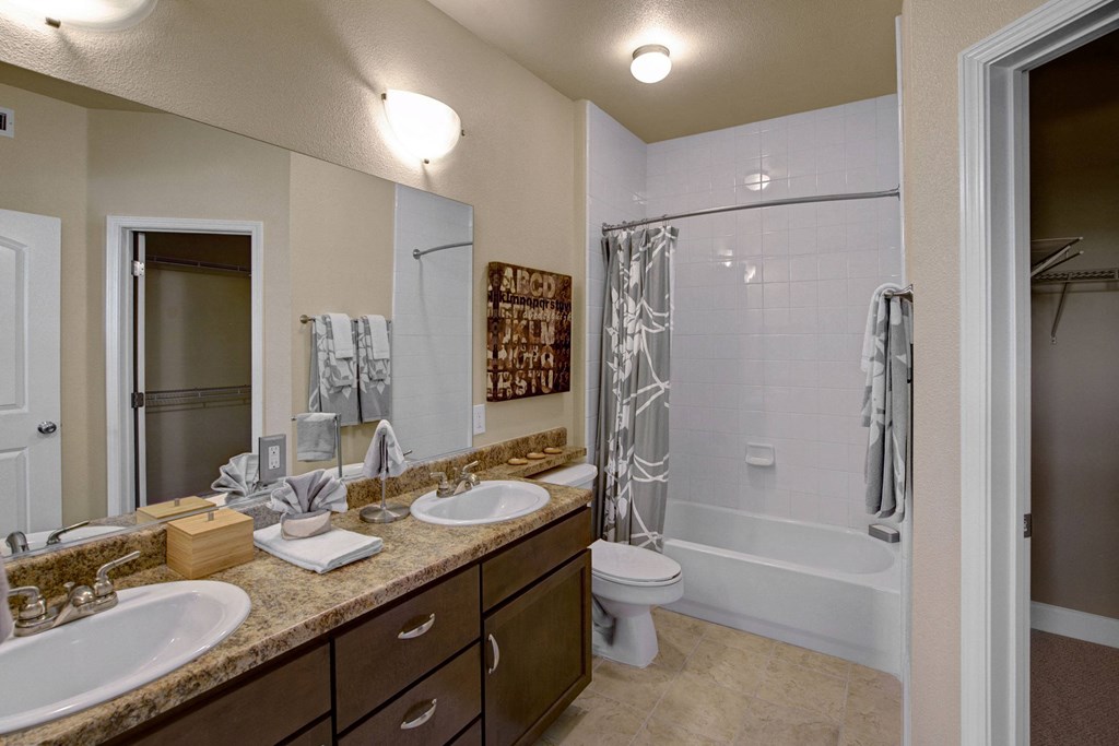 Modern bathroom here at Tuscany at Faudree Apartment Homes with double sinks on a granite countertop, large mirror, beige walls, tiled shower with a patterned curtain, and cozy lighting.