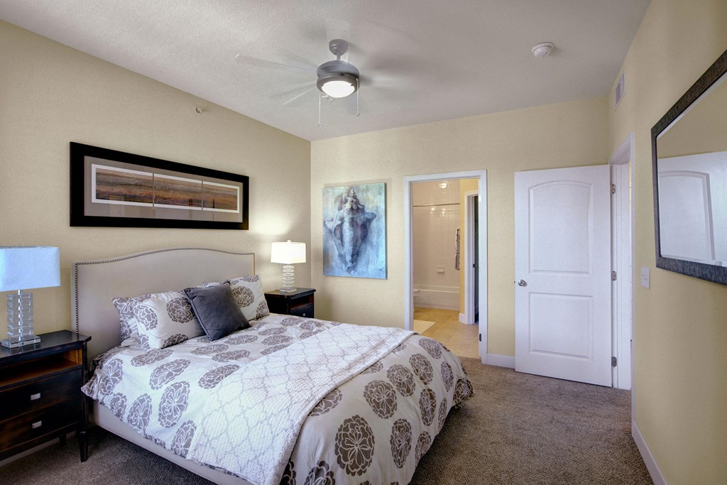 Modern bedroom here at Tuscany at Faudree Apartment Homes with soft lighting and neutral tones. Features a double bed with patterned bedding, nightstands with lamps, a ceiling fan, and wall art. Door to a bright bathroom.