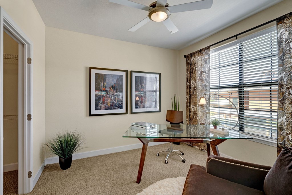 Modern home office here at Tuscany at Faudree Apartment Homes with a glass desk, brown leather chair, and a plant by the window. Two abstract art pieces on a beige wall. Bright and organized.