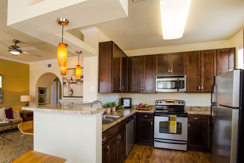 Stylish kitchen here at Tuscany at Faudree Apartment Homes with dark wood cabinets, stainless steel appliances, and granite countertops. Warm lighting and a cozy, inviting ambiance.