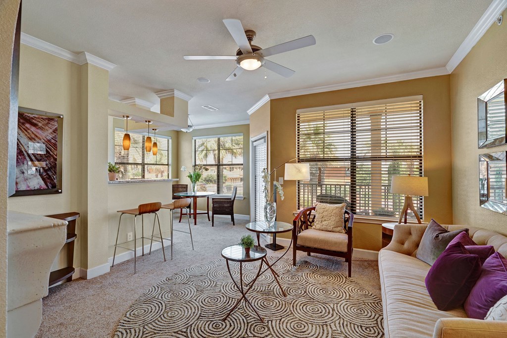 Stylish living room here at Tuscany at Faudree Apartment Homes with a beige sofa, purple cushions, circular rug, and glass table. Sunlit dining area with wicker chairs and pendant lights. Cozy ambiance.