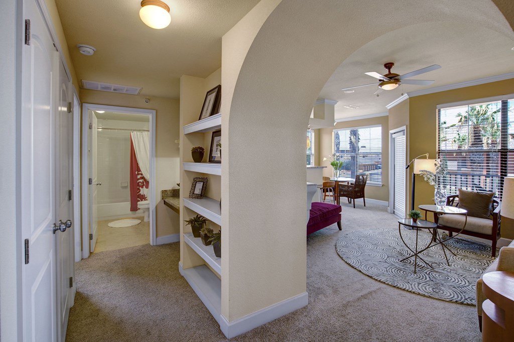 Spacious living area here at Tuscany at Faudree Apartment Homes with beige walls, archway, and carpeted floor. Features a circular rug, cozy seating, ceiling fan, and visible dining area. Warm, inviting tone.