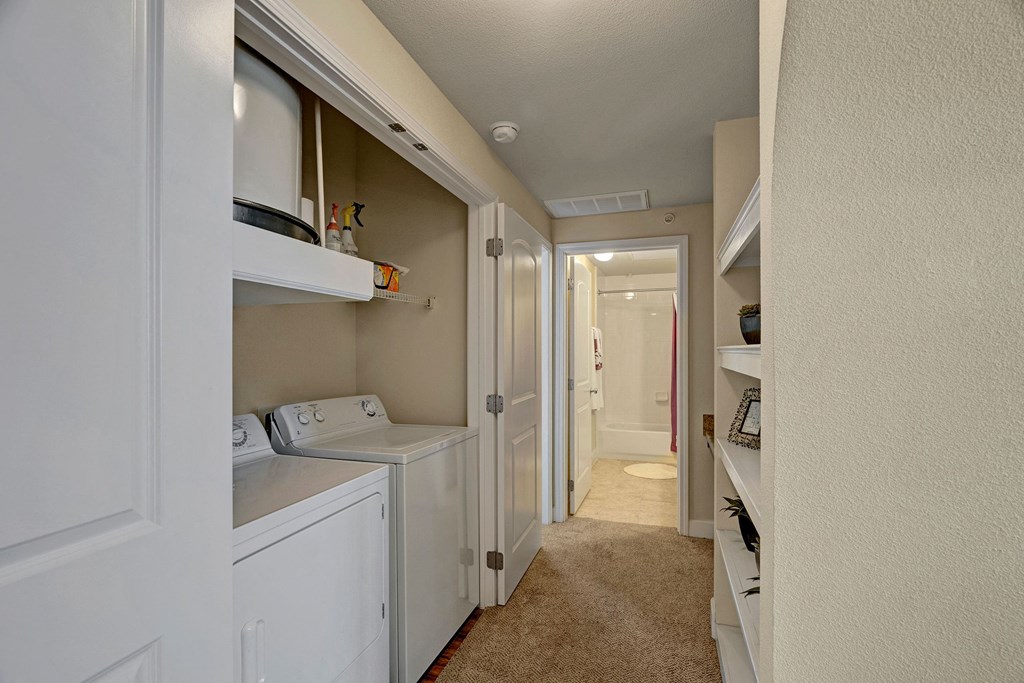 Narrow hallway here at Tuscany at Faudree Apartment Homes with beige walls and carpet. On the left, a laundry alcove with a washer and dryer; shelves on the right. A bathroom is visible at the end.