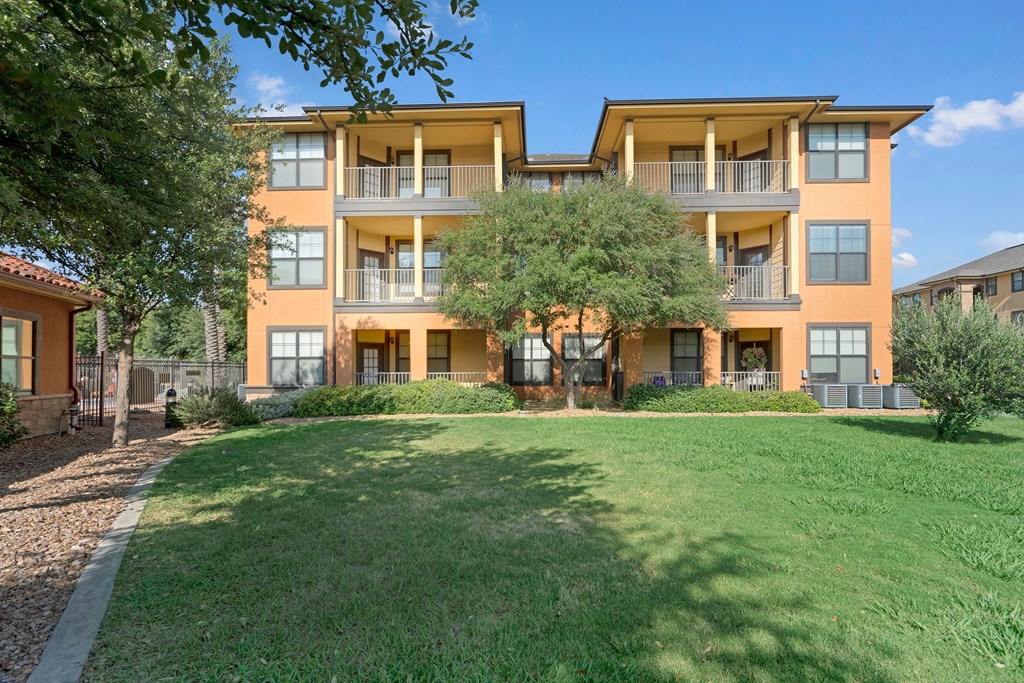 Three-story orange apartment building here at Tuscany at Faudree Apartment Homes with balconies and large windows, surrounded by lush green grass and trees under a clear blue sky.
