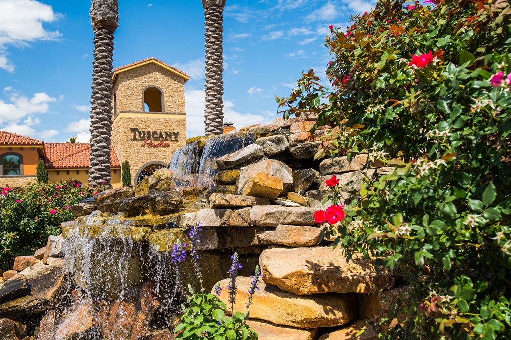 Stone waterfall surrounded by vibrant flowers and palm trees, with a building labeled "Tuscany" in the background under a bright blue sky.