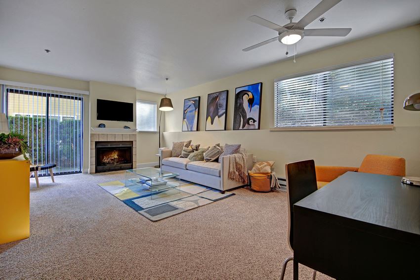 Living room with couch, desk, armchair, large windows, ceiling fan, fireplace, and sliding glass door out to a patio Foster Greens Living Room Apartments in Tukwila, WA
