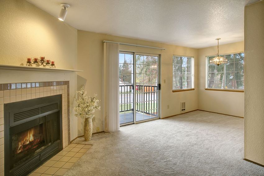 A spacious living room with a fireplace, a view into the dining area, and large sliding glass doors that lead out to the private balcony Heron View Living Room Apartments in Kenmore, WA - Photo Gallery