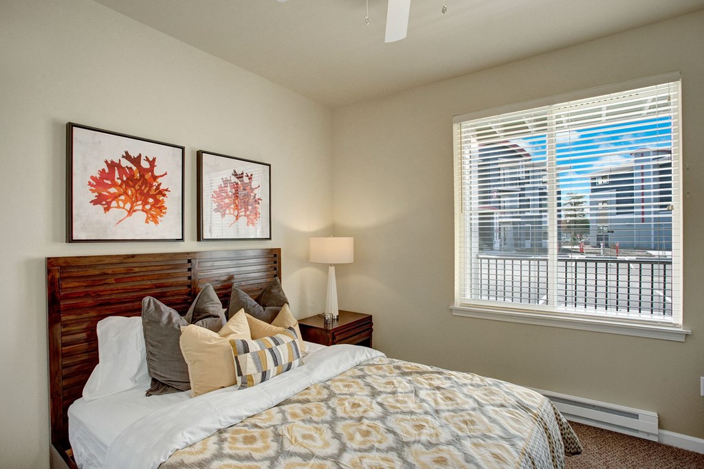 A bedroom with a large window looking out to the patio and the rest of the property with a bed, nightstand, and ceiling fan Solara Bedroom Apartments in Moses Lake, WA