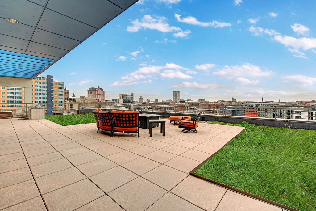 Rooftop patio at Avenir Apartments with modern seating, overlooking a cityscape. Surrounded by a low grassy area, under a blue sky with scattered clouds. Relaxing ambiance.